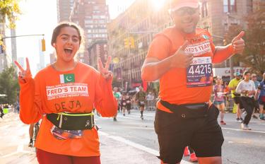  A woman and man, both wearing orange T-shirts with the words “Fred’s Team,” flash victory signs and thumbs-ups mid-race. 
