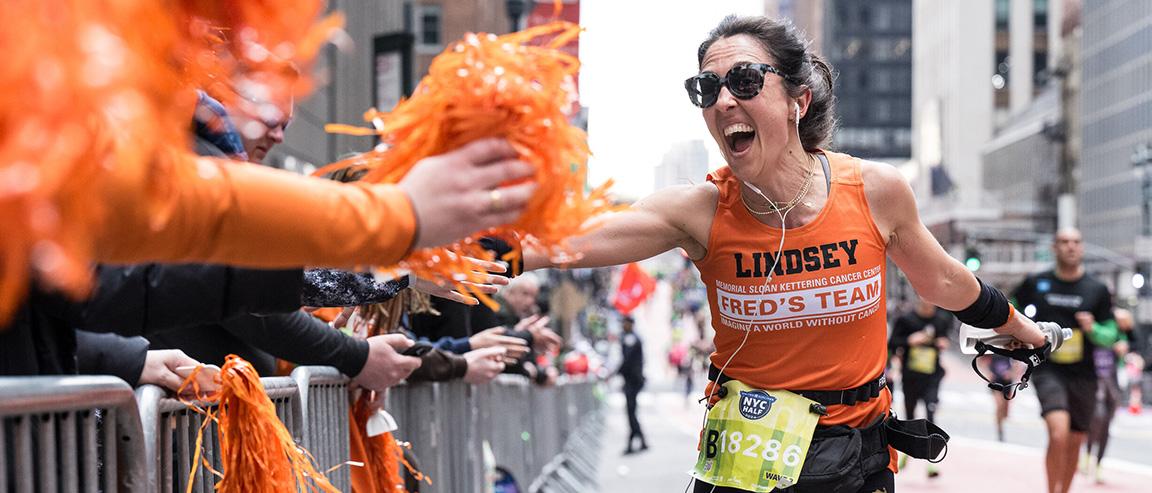 A runner with dark hair in a ponytail in an orange “Fred’s Team” tank top screams and holds out her right hand to a spectator mid-race as they raise money for cancer research. 