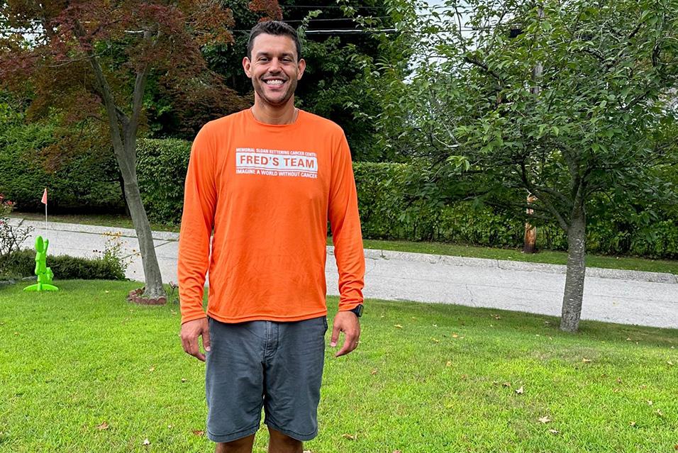 A person wearing a long-sleeved orange Fred’s Team shirt smiles while standing on a bright green lawn.  