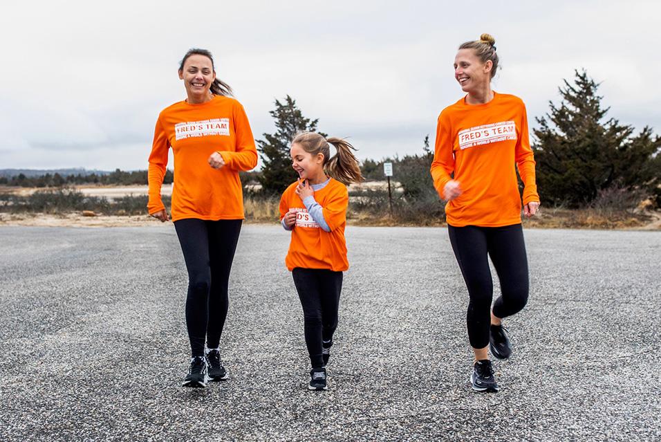 Three runners, one of them young, smile mid-stride. They all wear long-sleeved orange Fred’s Team shirts, black leggings, and black sneakers.  