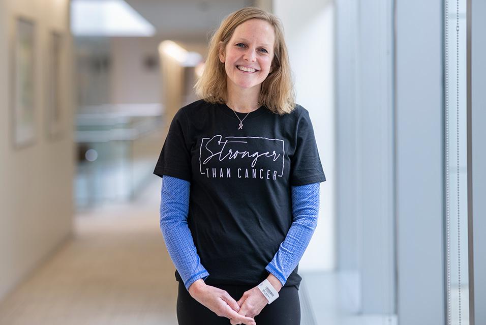 A person stands in a bright hallway, smiling. Her black T-shirt says “Stronger than cancer” on the front.  
