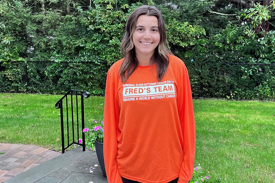 A person wearing a long-sleeved orange Fred’s Team shirt stands smiling in a yard with grass and trees in the background. 