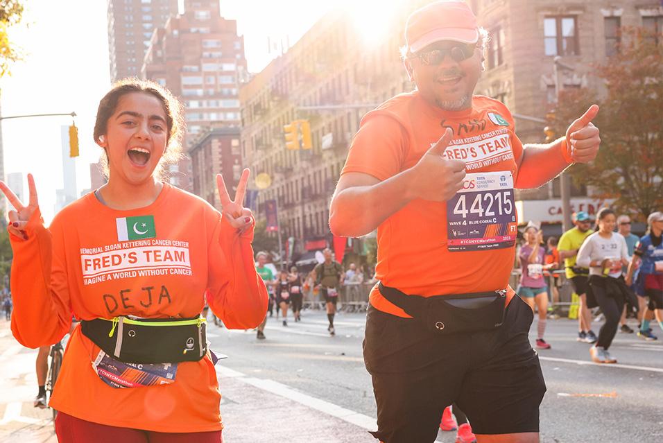 Two people, one wearing a hat and sunglasses, smile as they run in a race. Both are wearing orange Fred’s Team shirts. 