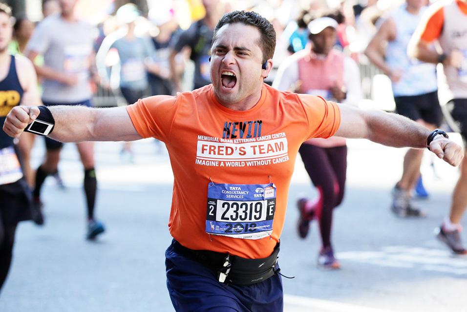 A man wearing an orange T-shirt with “Fred’s Team” and “Kevin” written on it flexes his arms mid-run, an victorious look on his face. 
