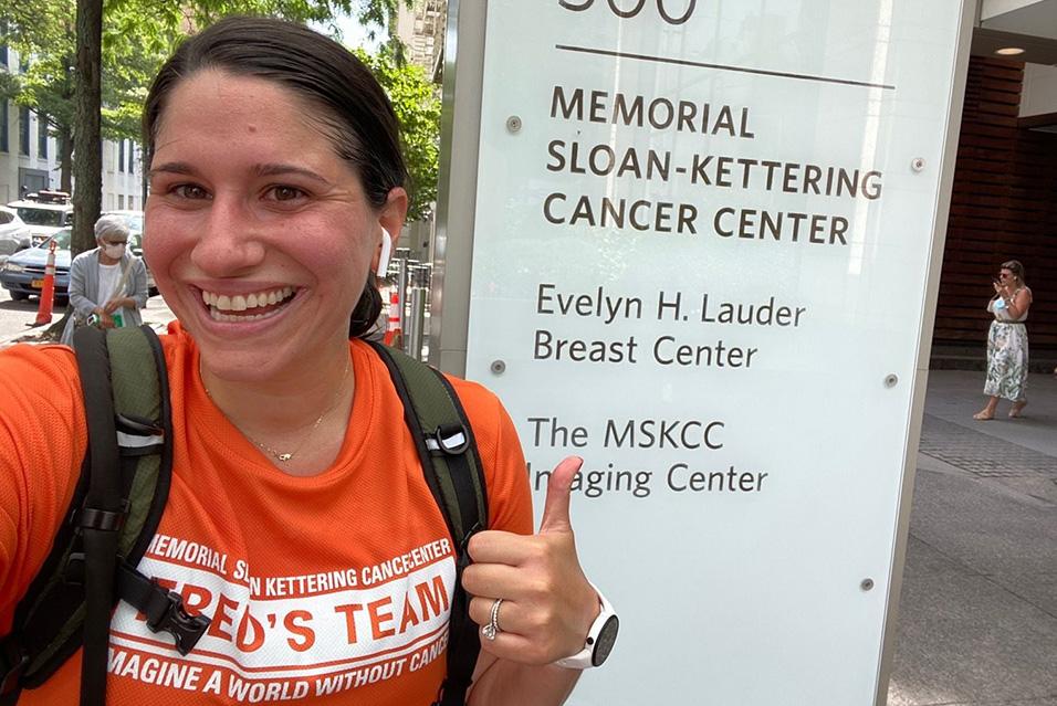 A woman wearing an orange Fred’s Team T-shirt gives a thumbs-up in front of a Memorial Sloan Kettering Cancer Center sign. 