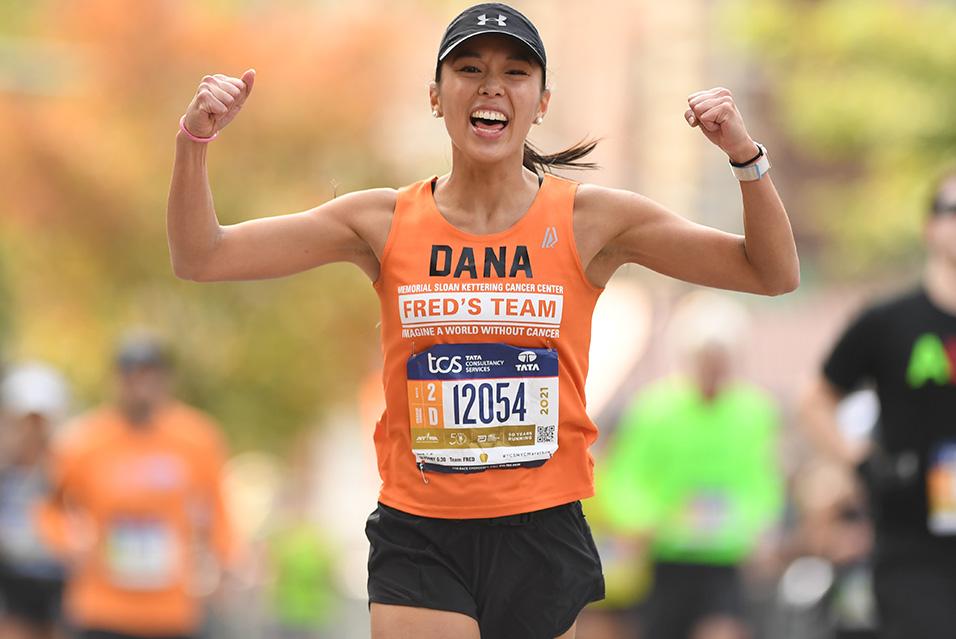 A woman in an orange tank top with “Fred’s Team” and “Dana” written on it runs with her fists in the air, wearing a joyful, proud smile. 