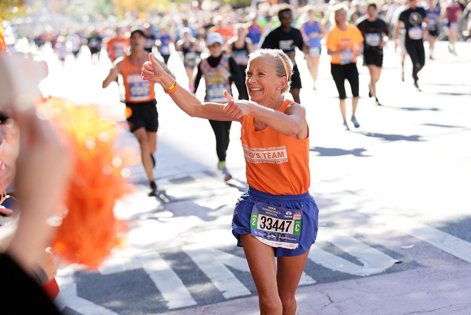 A woman smiles and gives two thumbs up while running a race. 