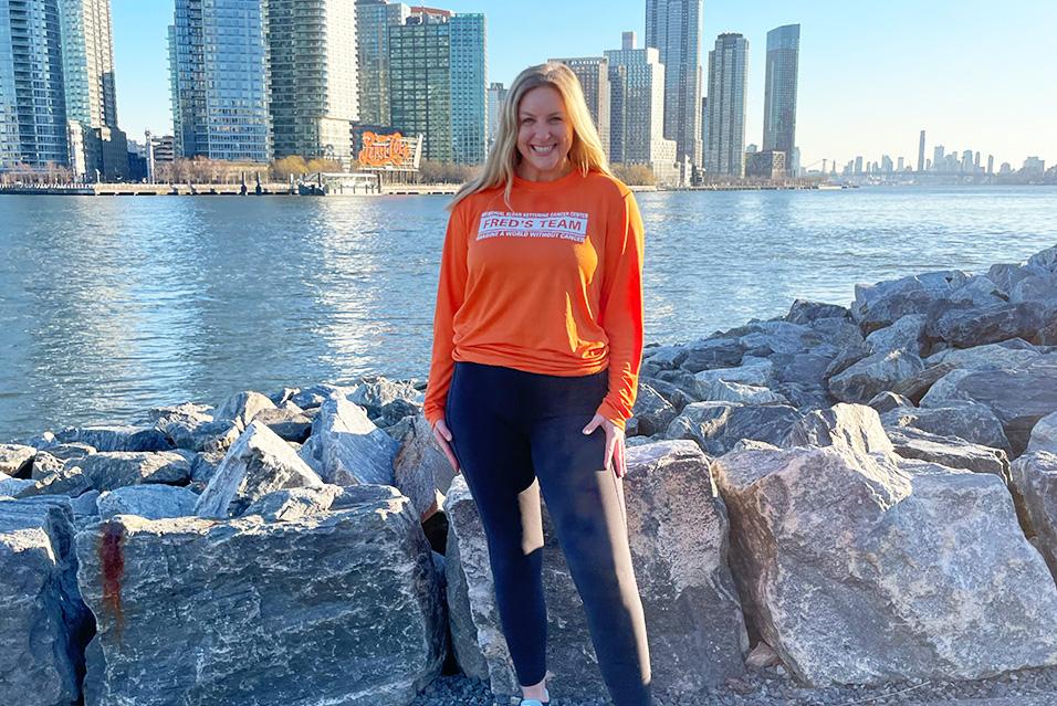 A woman wearing an orange Fred’s Team shirt stands smiling with a waterfront in the background. 