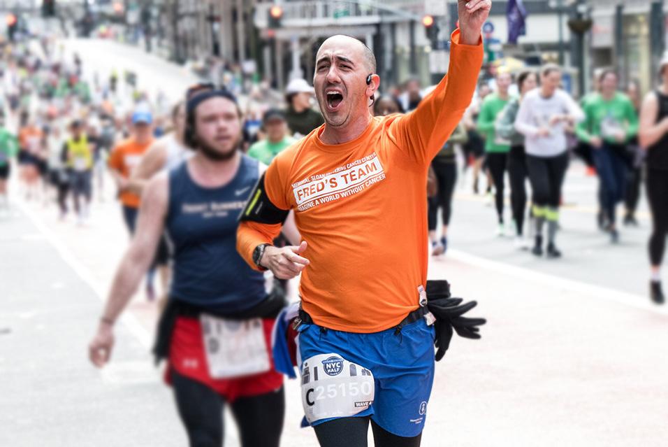 A runner with a shaved head wearing a long-sleeved orange Fred’s Team shirt cheers and waves to the crowd during a race. 