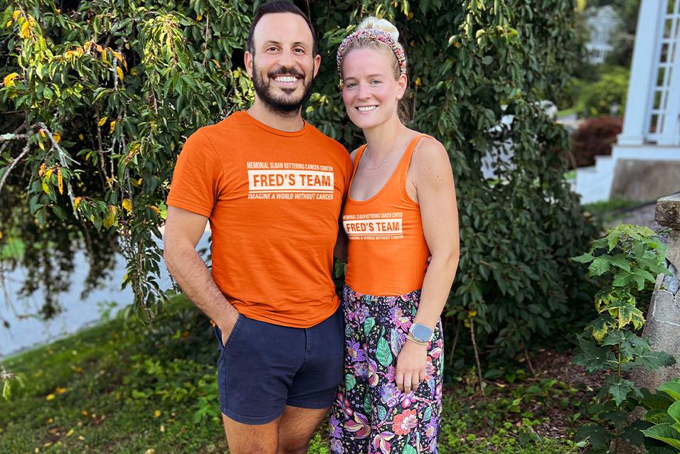 A man and woman stand side by side in front of a tree. They are both smiling and wearing orange Fred’s Team shirts. 